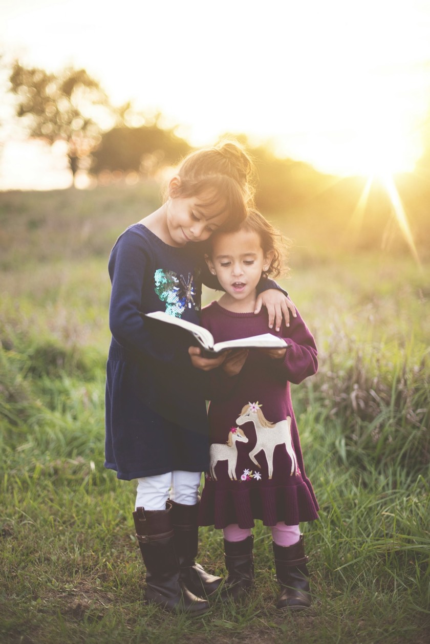 Two young sisters reading a book together outdoors in warm evening light